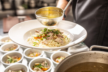 Chef in restaurant kitchen pours broth into a chicken soup with noodles, meat and vegetable