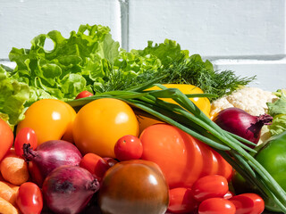 A pile of fresh, healthy and colorful vegetables in bright sunlight with white brick wall in background. Tomatoes, onions, carrots, peppers, cauliflower, salad, dill and greens