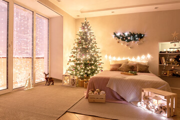 spacious bedroom with panoramic window with christmas tree near the bed. cozy New Year's decor of the room lit by the magic glow of garland lights. Festive atmosphere in the evening indoor