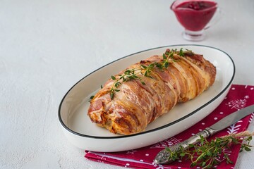 Cold appetizer, whole baked turkey and minced meat roll wrapped in bacon, on a white plate against a light concrete background.