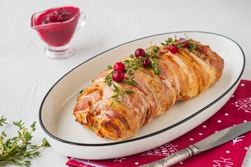 Cold appetizer, whole baked turkey and minced meat roll wrapped in bacon, on a white plate against a light concrete background.