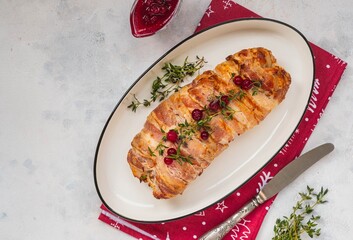 Cold appetizer, whole baked turkey and minced meat roll wrapped in bacon, on a white plate against a light concrete background.