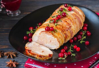 Cold appetizer, whole baked turkey and minced meat roll wrapped in bacon, on a black plate on a dark wooden background.