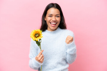 Young Colombian woman holding sunflower isolated on pink background celebrating a victory in winner position