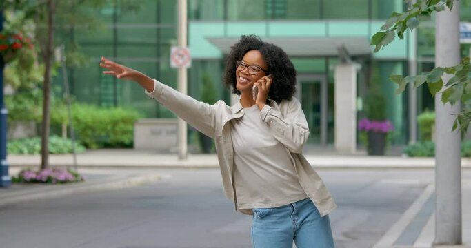 Attractive smiling African woman with curly hair calling taxi and having a conversation on Cell Phone on the street, dressed in a light shirt and blue jeans 