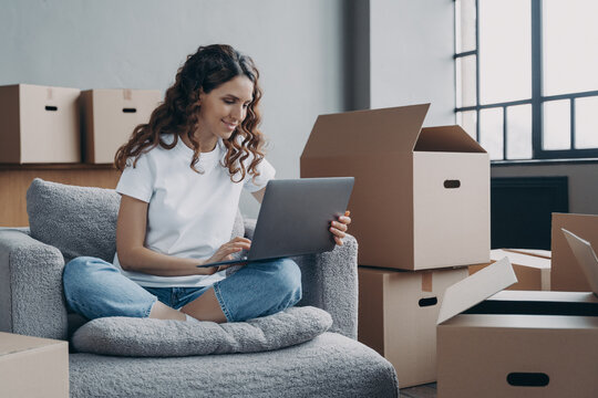 Smiling hispanic woman using laptop choosing moving service for moving in new home on relocation day