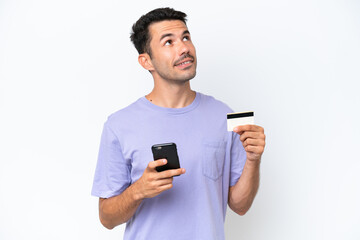 Young handsome man over isolated white background buying with the mobile with a credit card while thinking