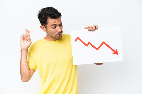 Young handsome man over isolated white background holding a sign with a decreasing statistics arrow symbol with fingers crossing