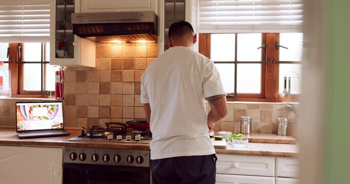 Man cooking food from recipe on laptop in his kitchen, cutting vegetables. Young guy alone using his computer to follow recipes for dinner or supper. Chef, cook and having a healthy meal at home