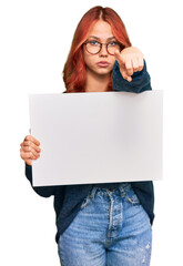 Young redhead woman holding blank empty banner pointing with finger to the camera and to you, confident gesture looking serious