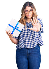 Young caucasian woman holding gift with open hand doing stop sign with serious and confident expression, defense gesture