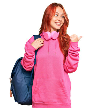 Young redhead woman holding student backpack and books pointing thumb up to the side smiling happy with open mouth