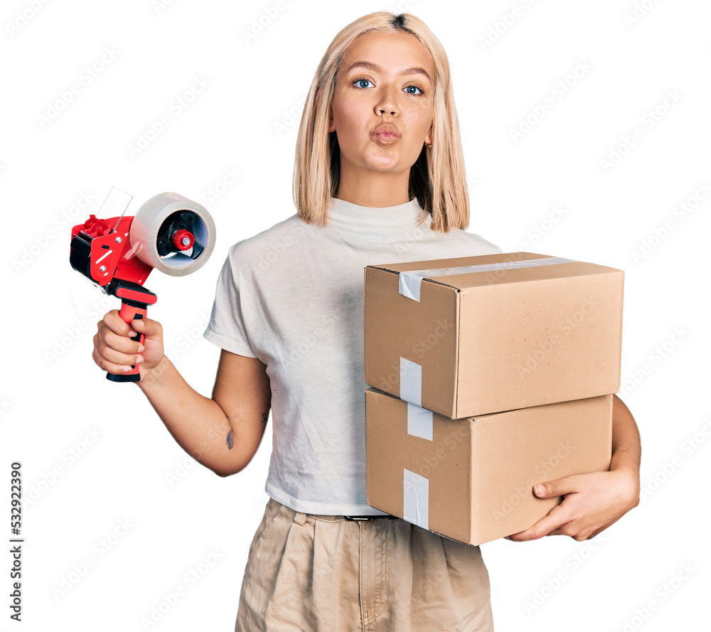 Poster beautiful young blonde woman holding packing tape and cardboard boxes looking at the camera blowing 