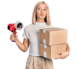 Beautiful young blonde woman holding packing tape and cardboard boxes looking at the camera blowing...