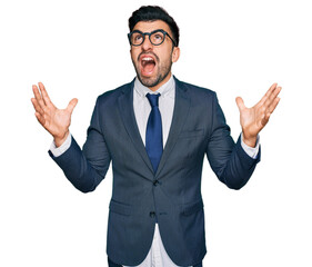 Hispanic man with beard wearing business suit and tie crazy and mad shouting and yelling with aggressive expression and arms raised. frustration concept.