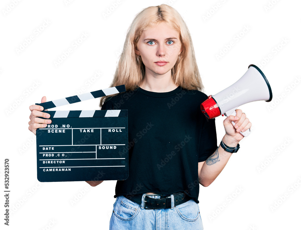 Wall mural Young caucasian woman holding video film clapboard and megaphone relaxed with serious expression on face. simple and natural looking at the camera.