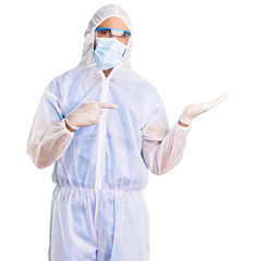 Young hispanic man wearing doctor protection coronavirus uniform and medical mask amazed and smiling to the camera while presenting with hand and pointing with finger.