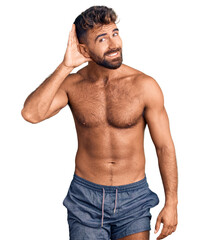 Fototapeta premium Young hispanic man wearing swimwear shirtless smiling with hand over ear listening an hearing to rumor or gossip. deafness concept.