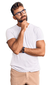 Young hispanic man wearing casual clothes and glasses with hand on chin thinking about question, pensive expression. smiling with thoughtful face. doubt concept.