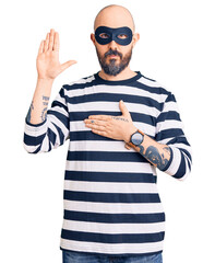 Young handsome man wearing burglar mask swearing with hand on chest and open palm, making a loyalty...