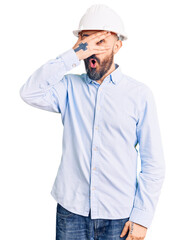 Young handsome man wearing architect hardhat peeking in shock covering face and eyes with hand, looking through fingers with embarrassed expression.