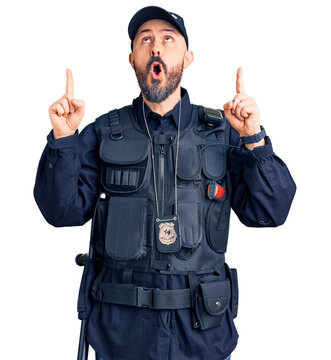 Young handsome man wearing police uniform amazed and surprised looking up and pointing with fingers and raised arms.