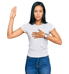 Beautiful hispanic woman wearing casual white tshirt swearing with hand on chest and open palm, making a loyalty promise oath