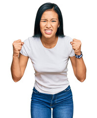 Beautiful hispanic woman wearing casual white tshirt angry and mad raising fists frustrated and...