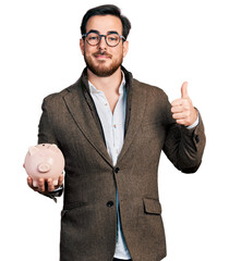 Young hispanic man holding piggy bank smiling happy and positive, thumb up doing excellent and...