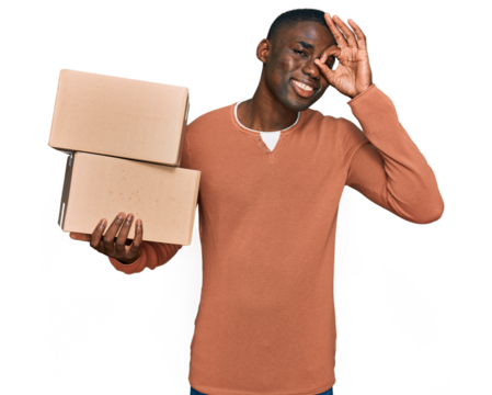 Young african american man holding delivery package smiling happy doing ok sign with hand on eye looking through fingers