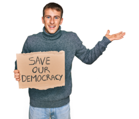 Young blond man holding save our democracy protest banner celebrating victory with happy smile and winner expression with raised hands