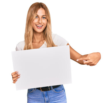 Beautiful blonde young woman holding blank empty banner smiling happy pointing with hand and finger
