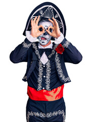 Young man wearing day of the dead costume over background doing ok gesture like binoculars sticking tongue out, eyes looking through fingers. crazy expression.