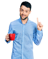 Hispanic man with beard drinking a cup coffee smiling with an idea or question pointing finger with...