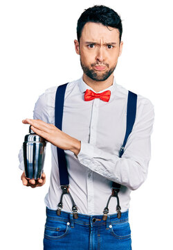Hispanic man with beard preparing cocktail mixing drink with shaker puffing cheeks with funny face. mouth inflated with air, catching air.