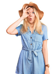 Young beautiful blonde woman wearing summer hat and dress doing ok gesture shocked with surprised face, eye looking through fingers. unbelieving expression.