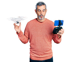 Fototapeta na wymiar Middle age hispanic man with grey hair using drone with remote control making fish face with mouth and squinting eyes, crazy and comical.