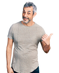 Middle age hispanic with grey hair wearing casual grey t shirt smiling with happy face looking and pointing to the side with thumb up.