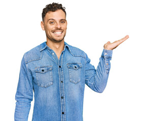 Young hispanic man wearing casual denim jacket smiling cheerful presenting and pointing with palm of hand looking at the camera.