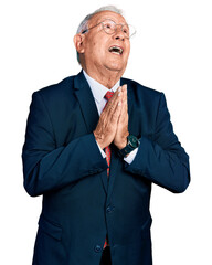 Senior man with grey hair wearing business suit and glasses begging and praying with hands together...