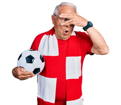 Senior man with grey hair football hooligan holding ball peeking in shock covering face and eyes with hand, looking through fingers with embarrassed expression.