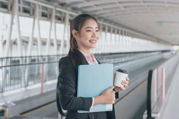 young Asian businesswoman in black suit holding folder and cup of coffee and standing on walkway station.