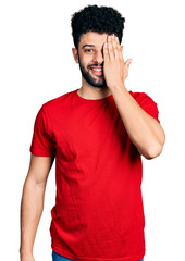 Fototapeta na wymiar Young arab man with beard wearing casual red t shirt covering one eye with hand, confident smile on face and surprise emotion.