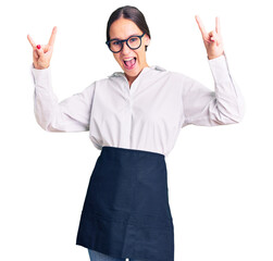 Beautiful brunette young woman wearing professional waitress apron shouting with crazy expression doing rock symbol with hands up. music star. heavy music concept.