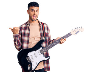 Young hispanic man playing electric guitar pointing thumb up to the side smiling happy with open mouth