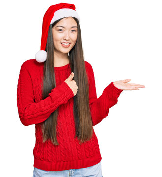 Young chinese woman wearing christmas hat showing palm hand and doing ok gesture with thumbs up, smiling happy and cheerful