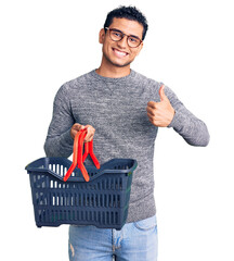 Hispanic handsome young man holding supermarket shopping basket smiling happy and positive, thumb...