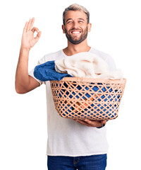 Young handsome blond man holding laundry basket with clothes doing ok sign with fingers, smiling...