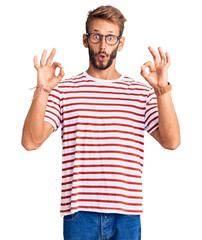 Handsome blond man with beard wearing casual clothes and glasses looking surprised and shocked doing ok approval symbol with fingers. crazy expression