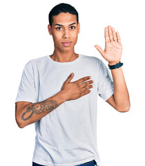 Young hispanic man wearing casual white t shirt swearing with hand on chest and open palm, making a...